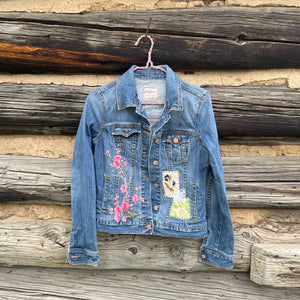Denim jacket with floral and abstract designs hanging against a wooden wall.
