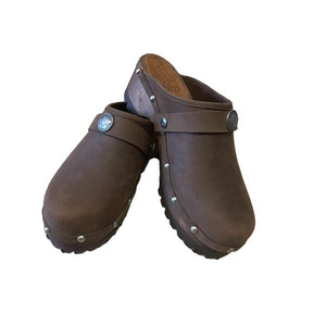 A pair of brown leather clogs with pewter metal studs and a decorative pewter buffalo medallion on the strap, shown on a plain white background.