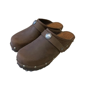 A pair of brown leather clogs with pewter metal studs and a decorative pewter buffalo medallion on the strap, shown on a plain white background.