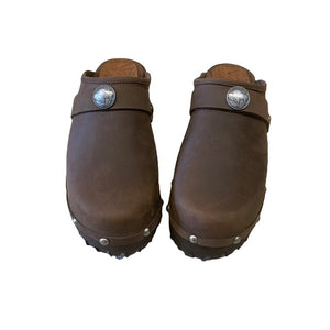 A pair of brown leather clogs with pewter metal studs and a decorative pewter buffalo medallion on the strap, shown on a plain white background.