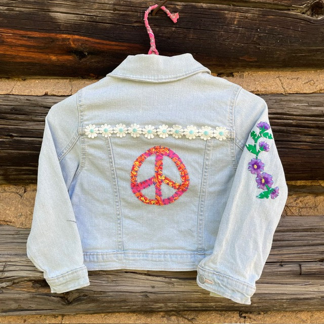 Back of jean jacket with large peace sign patch and daisy chain patch on back and a flower patch on the right arm.