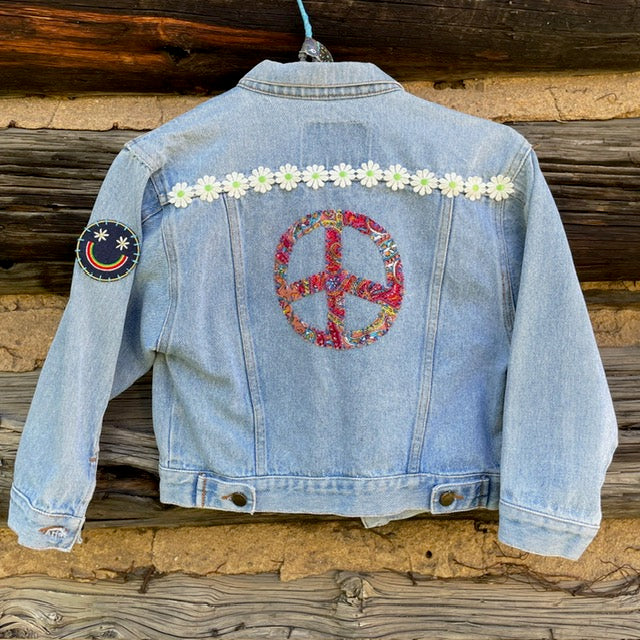 Back of kids jean jacket with large peace sign, daisy chain, and blue smiley face patch on left shoulder.