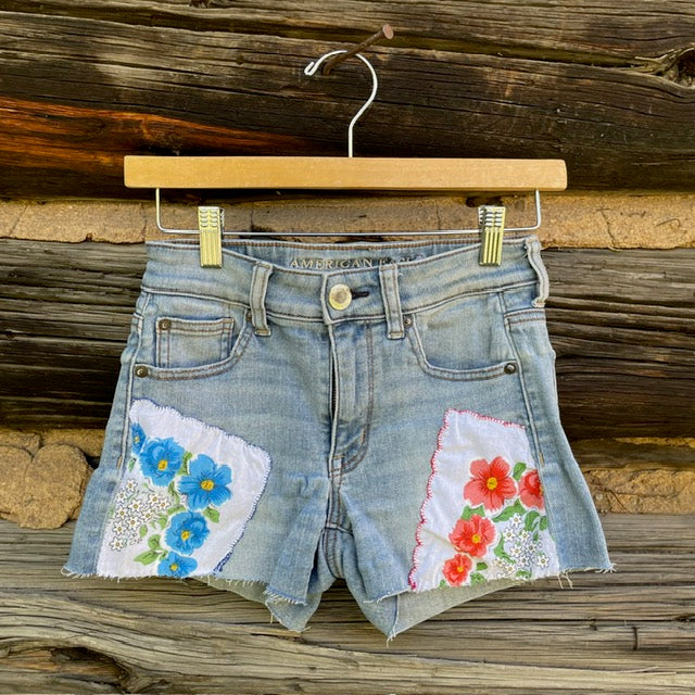 Front of jean shorts with a blue lowered napkin patch anda red flowered napkin patch over the front pockets.